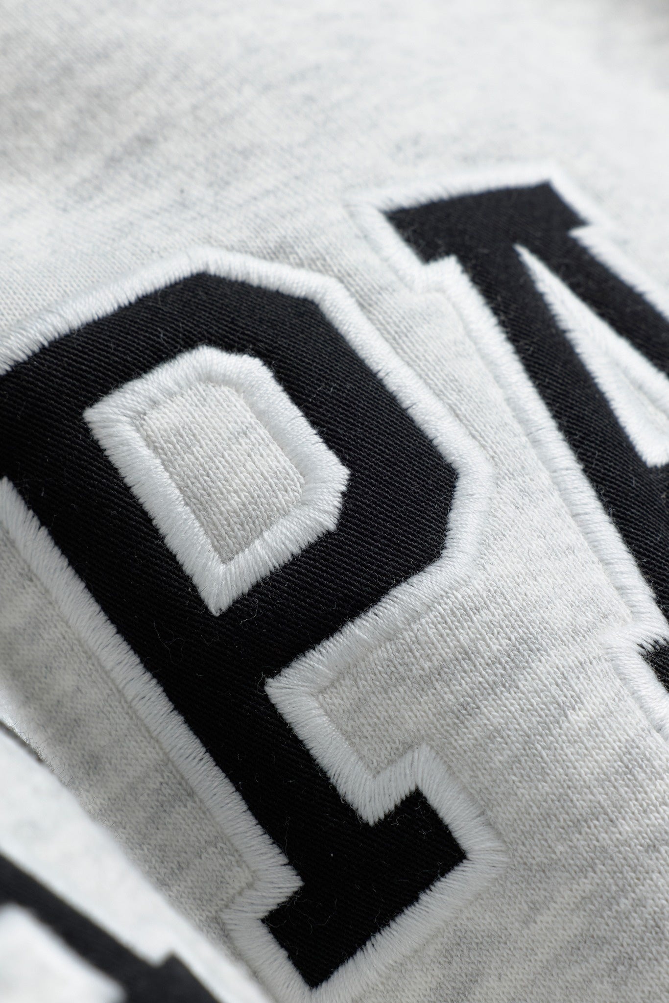 Close-up of black and white embroidered varsity lettering on a grey melange CASPARA crewneck sweatshirt.